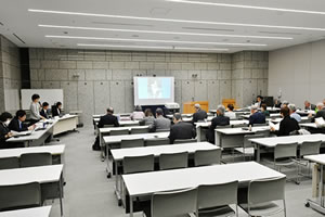 広い部屋、会場正面にはスクリーンが立てられ、何か投映されている。前方に向けて並べられた長机に東友会の参加者が着席している。側面壁際に、東友会の参加者に向けて並べられた長机があり、東京都の参加者が着席している。うち一人は立ってマイクを使い話している。