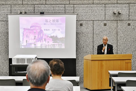 スクリーンに、原爆ドームの写真と「私と原爆」などの文字が投影されている。横に置かれた演台で、木村一茂さんがマイクを使い話している。