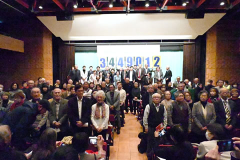 ほとんどの参加者が着席してカメラの方を向いている集合写真。奥の壇上で立っている人たちもいる。「3449012」の数字が大きく横書きされた紙が、壇上の高くから下げられている。