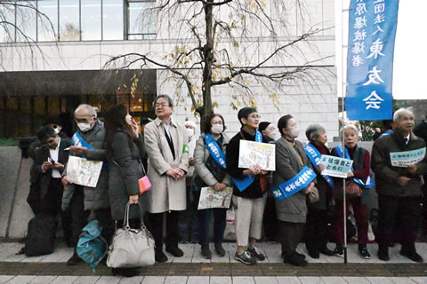 タスキを掛けた被爆者をはじめとした参加者たちが道に立っている。被爆者のひとりは、東友会ののぼりを立てている。「#被爆者とともに」のハッシュタグが大きく書かれた紙などを持っている人もいる。