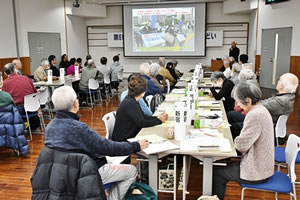 広い部屋、前方の天井からさげられたスクリーンに、なにか写真が投映されている。脇の演台で、マイクを持って立ち話している人がいる。並べられた長机に着席した参加者は、スクリーンや手元の資料を見ながら話を聞いている。