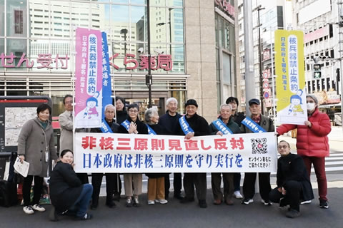 駅前歩道で、行動参加者の集合写真。タスキを掛けた被爆者や他の参加者が立って横に並び、「非核三原則見直し反対 日本政府は非核三原則を守り実行を」と大きく書かれた横断幕を広げ持っている。「核兵器禁止条約 日本政府も署名・批准を」と書かれたのぼりを持っている人もいる。