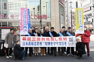 駅前歩道で、行動参加者の集合写真。タスキを掛けた被爆者や他の参加者が立って横に並び、「非核三原則見直し反対 日本政府は非核三原則を守り実行を」と大きく書かれた横断幕を広げ持っている。「核兵器禁止条約 日本政府も署名・批准を」と書かれたのぼりを持っている人もいる。