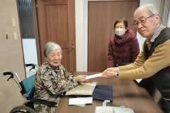 訪問先の車いすの方に、訪問者の一人が見舞金を手渡している。奥にもう一人訪問者が写っている。3名ともカメラに笑顔を向けている。