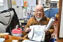 椅子に座った訪問先の方が、受け取った見舞金などを旨の前に掲げ持ち、カメラに笑顔を向けている。