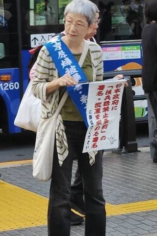 街頭で、青いたすきを掛けて署名板を持つ人。署名板には、通行人から見えるよう「日本政府に『核兵器禁止条約』の署名・批准を求める署名」と書かれたk紙がさげられている。
