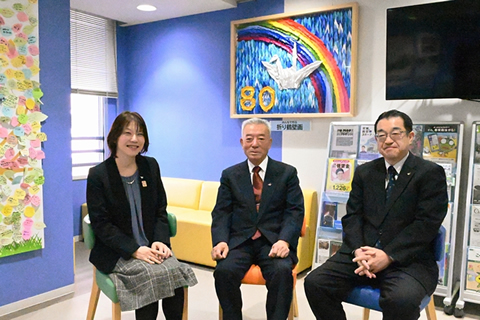 椅子に座ってカメラに笑顔を向けている3人。背後の壁に、無数の折り鶴で作られた大きいモザイクがかけられている。モザイクは、中央に折り鶴、その背景に虹のかかった空、左下に「80」の文字が置かれている。