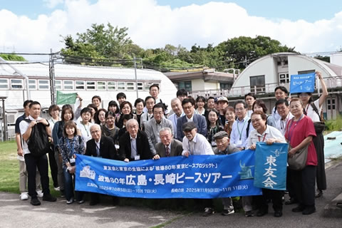 ツアー参加者の集合写真。最前列の人たちが、「広島・長崎ピースツアー」と書かれた横断幕を広げ持っている。建物前の広い庭のような場所、舗装された通路で。