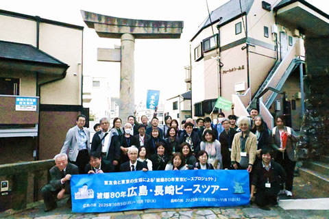 s狭い道で、ツアー参加者の集合写真。最前列の人たちが、「広島・長崎ピースツアー」と書かれた横断幕を広げ持っている。鳥居は、高さのある民家に埋もれるような様相。