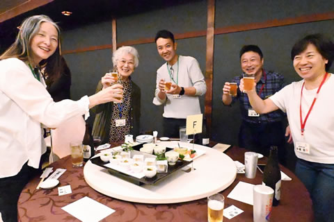 丸いテーブルを囲んで立ち、ビールの注がれたグラスを持ってカメラに笑顔を向ける参加者たち。