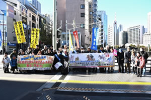 参加者が、歩道の端に横に広がって並んでいる。東友会関係の参加者はたすきをかけている。最前列の人たちは、馬のイラストも描かれた横断幕(写真では文字がつぶれてしまい読めない)、「高校生署名 日本政府は核兵器禁止条約に署名・批准を！」などと書かれた横断幕を広げ持っている。東友会を始め、団体ののぼりも立てられている。のぼりには「核兵器廃絶 子どもを守ろう」と書かれたものもある。