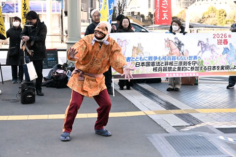 手ぬぐいの頬かむりをして面を着け、着物のような衣装を着た人が、横断幕を持った参加者たちを背にして歩道で踊っている。