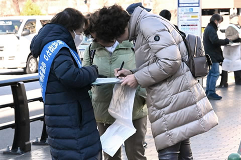 東友会のタスキをかけた参加者が持つ署名板で、1人の通行人がペンを持ち署名をしている。その連れと思われる人が、一緒に署名用紙を見ている。