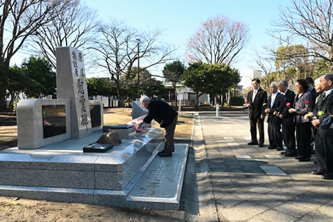 役員1名が慰霊碑に献花している。その後ろ、慰霊碑から数歩さがったあたりに他の役員が花束を持つなどして並び、見守っている。