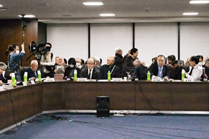 並べられた机に着席する参加者ら。それぞれの参加者の手元には紙の資料とペットボトルのお茶がおかれている。また、最前列の人の前には、向き合う席などから見えるように名札が置かれている。がある。テレビ局のものと思われる大型のビデオカメラが据えられ、操作している人がおり、隣には長い棒につけられたマイクを持っている人もいる