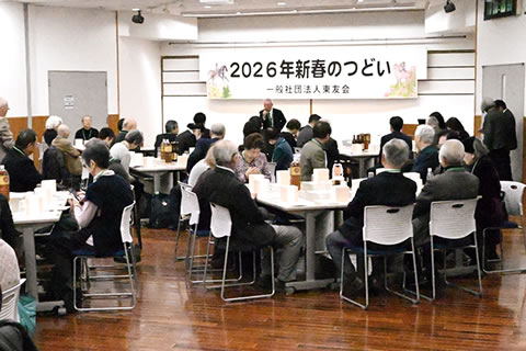 開場前方の壁天井近くに、「2026年新春のつどい 一般社団法人東友会」と大きく横書きされた紙がさげられている。文字を挟んで両端に、干支である馬のイラストもある。その手前で、マイクを持って参加者に向かって立って話している人が1人いる。参加者はテーブルの席について聞いている。