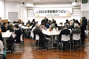 場前方の壁天井近くに、「2026年新春のつどい 一般社団法人東友会」と大きく横書きされた紙がさげられている。文字を挟んで両端に、干支である馬のイラストもある。その手前で、マイクを持って参加者に向かって立って話している人が1人いる。参加者はテーブルの席について聞いている。