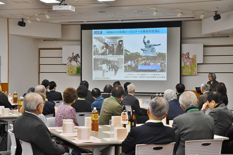 会場前方にさげられた大きいスクリーンに、写真三枚と説明の文章のある画像が投影されている。参加者たちが席に座ってそれを見ている。