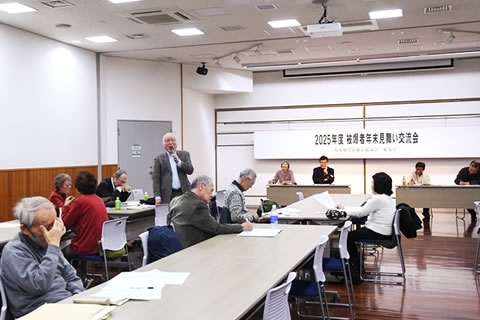 参加者の一人が席で立ち、マイクを使って話している。他の参加者たちは並べられたテーブルの席に着席し、メモを取るなどしながら聴いている。