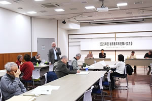 参加者の一人が席で立ち、マイクを使って話している。他の参加者たちは並べられたテーブルの席に着席し、メモを取るなどしながら聴いている。