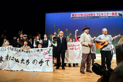 写っているのは舞台の一部。ギターを弾いている人や「高校生平和集会 in 焼津 未来は私たちの手で」と書かれた横断幕を広げ持つ高校生たちなどがいる。舞台奥の天井から、「被災72年2026年3・1ビキニデー集会」と大きく横書きされた看板がさげられている。