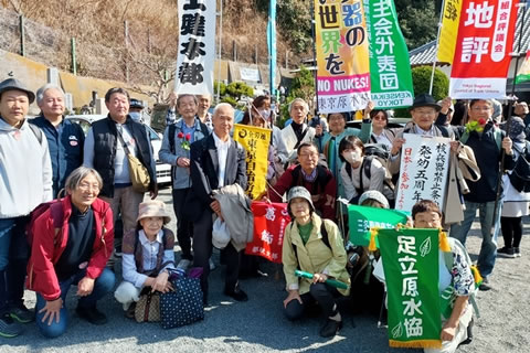 墓参行進に参加した人たちのうち20名ほどが集まってカメラの方を向いている集合写真。様々な団体の旗などを持っている人もいるが、立てられた旗は下の方の一部しか写っていない。また、小さい旗などを持っている人もいるが、文字は小さくて読めない。読めるのは「足立原水協」と、「核兵器禁止条約 発効五周年 日本も参加しよう」と書かれたもののみ。（それぞれ別の人が体の前に掲げている。）