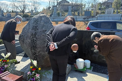 「原爆被害者の墓」の、墓石の背後にある地下納骨室のふたが開けられている。1人の人が納骨室に降り、上半身をだして、骨壺の一つを両手で持ち上げている。そばにもう一つ骨壺が置かれている。その様子を3人が近くで見守っており、1人は骨壺に向かって手を合わせている。墓石の間には花が供えられている。