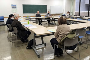 長方形に並べられた長机に、向き合う形で着席する参加者たち。机には紙の資料が配られている。