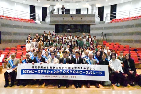 会場の客席に上に集まった参加者たち。最前列の椅子に座った人たちが、「核兵器廃絶と戦争のない平和な世界をめざして 2026ピースアクション in TOKYO ＆ ピースパレード」と書かれた横断幕を広げ持っている。被爆左yはタスキを掛けている。手前側の人たちは客席に座っており、奥側の人たちは顔が隠れないように立っている。