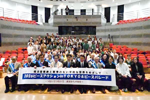 会場の客席に上に集まった参加者たち。最前列の椅子に座った人たちが、「核兵器廃絶と戦争のない平和な世界をめざして 2026ピースアクション in TOKYO ＆ ピースパレード」と書かれた横断幕を広げ持っている。被爆左yはタスキを掛けている。手前側の人たちは客席に座っており、奥側の人たちは顔が隠れないように立っている。