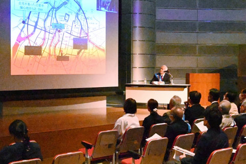 舞台奥の大きいスクリーンに、広島市の地図が投影されている。地図には爆心の位置と、爆心を中心とした円（半径は写真では不鮮明で読み取れない）などが描かれている。そのわきの長机で、木村一茂さんが椅子に座って話をしている。