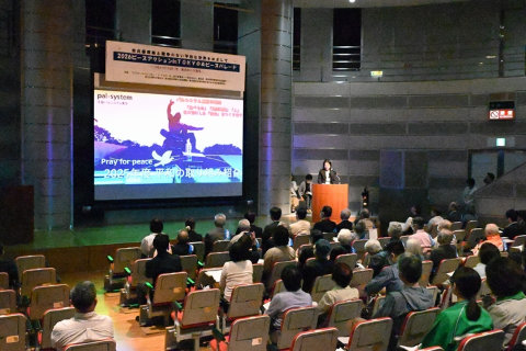 舞台の上のスクリーンに、長崎平和祈念像の写真またはイラストとなんらかの文章が投影されている。その脇の演台で、立って話をしている人がいる。