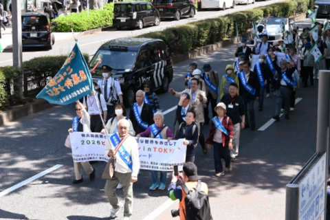 旗竿を持つ、タスキを掛けた被爆者を先頭に車道を歩くパレード参加者。旗竿を持つ被爆者の直後を歩く、タスキを掛けた被爆者含む数人が、横断幕（不鮮明かつ隠れているため、「ピースパレード」の他に書かれている内容は不明）を広げ持っている。その後ろに参加者の列が続いている。