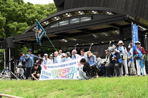 第五福竜丸のエンジンが展示されている屋根の下、タスキを掛けた東友会の参加者たちが、「国民平和大行進」広げ持っている。その脇で、1人がマイクを使い話している。横断幕のそばには、参加団愛の広報担当なのか、しゃがみこんで様子を撮影している人がいる。