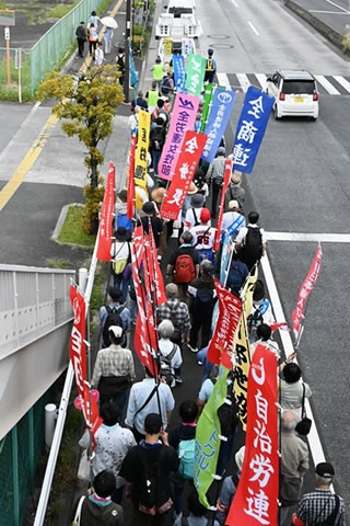 行進の先頭部分を後方から、歩道橋の上から見下ろして移した写真。行進の隊列は車道の限られた幅ぎゅうぎゅうになっているように見える。参加者の持つ、団体名などの書かれたのぼりが多数写っているが、風になびいていた文字が小さく写っていたりして、読めるものはわずか。（確実に読めるのは「全商連」「自治労連」の2つだけ。）