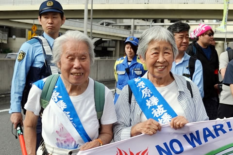 笑顔で、タスキを掛けて横断幕を持つた四辻さん片田さんの2人。(横断幕は上端のみ写っており、読めるのは「NO WAR！」のみ。)2人の背後に、行進を見張る警官が写っている。