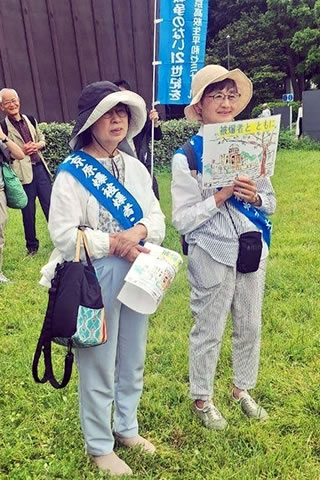 「東友会の青いたすきをかけた二人が、第五福竜丸展示館を背景に立っている。一人は「被爆者とともに」の文字も書かれた、原爆ドームのイラストを胸の前に掲げている。