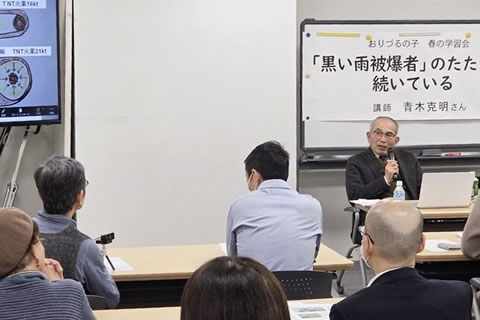 マイクを使って話す講師と、長机に座ってそれを聴く参加者たち。講師の背後のホワイトボードに、「『黒い雨被爆者』のたたかいは続いている」と大きく書かれた紙が張られている。(ただし、文字が一部写真に入りきっていない)また、大型モニターに何か資料の画像が映し出されているが、モニターのごく一部しか写真に入っていないため詳細はわからない。
