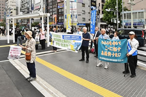 署名判を持って立ち、通行人に訴える人の背後に、横断幕を広げもつ人たちや、東友会ののぼりを持っている人、「われら生命もて ここに証す 原爆許すまじ」と書かれた東友会の旗を広げもつ被爆者らいる。横断幕には、「日本政府は核兵器禁止条約に参加・批准を！」など書かれている。