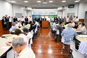 会場前方、「第78回 一般社団法人東友会 第70回 東京都原爆被害者協議会 定期総会」と大きく横書きされた看板が天井近くに下げられている。その下には東友会の旗が下げられている。その手前に、選出された役員が一列の横並びで立っている。他の出席者は、並べられた長机に着席し、役員たちのほうを見ていたり、資料に目を落とすなどしている。