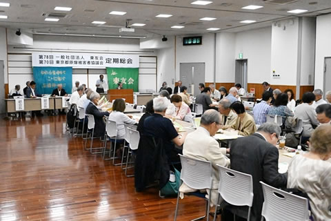 「第78回 一般社団法人東友会 第70回 東京都原爆被害者協議会 定期総会」と大きく横書きされた看板が前方の天井近くから下げらた会場、演台でマイクを使い話している人がいる。他の出席者たちは席に着き、資料に目を落とすなどしながらそれを聴いている。