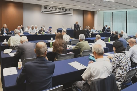 会場前方、天井から「第71回定期総会」と大きく横書きされた看板がさげられている。演台に立った1人がマイクを使って話している。他の参加者たちが、並べられた机に着席し、手元の資料に目を落とすなどしながら聴いている。