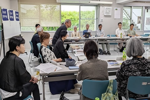 長方形に並べられた長机に、向き合う形で着席する参加者たち。机には紙の資料が配られている。1人が席で立って何か発言しており、他の参加者たちがそれを聞いている。