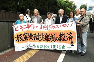 「ビキニ事件はまだ終わっていない！」「核実験被災船員の救済を」「ビキニ労災訴訟を支援する会」と書かれた横断幕を路上で広げ持つ、原告、弁護士、支援者たち。