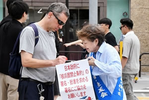 たすきをかけた被爆者の持つ署名板で、被爆者より頭一つ分背が高い、外国にルーツがあると思われる人が署名している。被爆者は、書きやすいよう署名板を自分の胸の上の高さまで持ち上げて支えている。署名板には、「日本政府に『核兵器禁止条約』の署名・批准を求める署名にご協力をお願いします」と書かれた紙が下げられている。