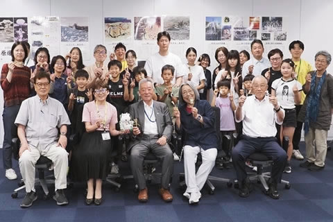 大人子どもあわせ50人ほどの参加者の集合写真。みなカメラに向けて笑顔で、ピースサインをしている人もいる。最前列の人たちは椅子に座っている。背後の壁には原爆展パネルが掲示されている。