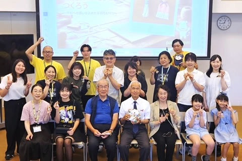 大人子どもあわせ30人ほどの参加者の集合写真。みなカメラに向けて笑顔で、ピースサインをしている人もいる。最前列の人たちは椅子に座っている。背後に天井から下げられた大きいスクリーンがあり、「せんそうほうき」の完成品が映写されている。