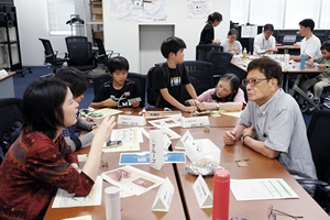 テーブルをはさんで、何か会話している人たち。同じテーブルの奥では、親子が作業している。テーブルの上には、「せんそうほうき」の作り方など書かれていると思われる紙や、材料などが広げられている。