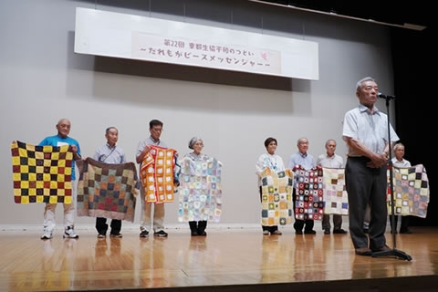 舞台の奥側に、手編みのひざ掛けを客席に向け広げ持って並ぶ被爆者たちがいる。舞台手前のスタンドマイクを使って、家島昌志代表理事が挨拶している。