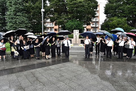 慰霊碑の手前に、慰霊碑を挟む形で並ぶ献花式参加者たち。みな傘をさして立っている。